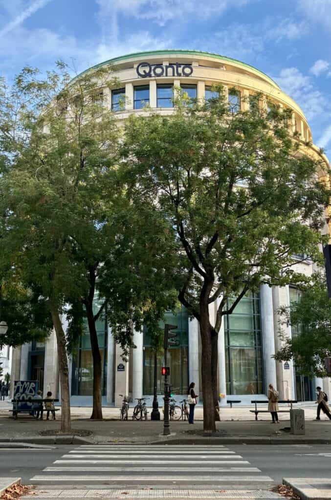 Façade arrondie de l’immeuble Qonto à Paris, logo en haut, arbres au premier plan et passage piéton