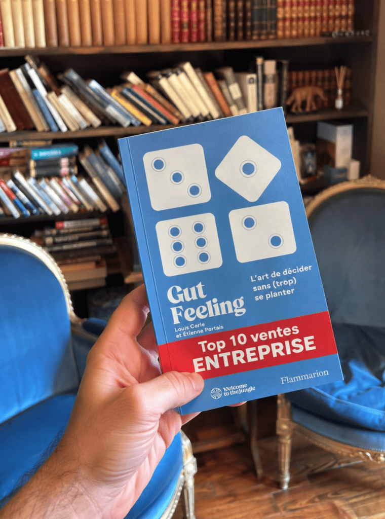 Une main tient le livre Gut Feeling de Louis Carle et Étienne Portais, bandeau Top 10 ventes Entreprise, devant une bibliothèque