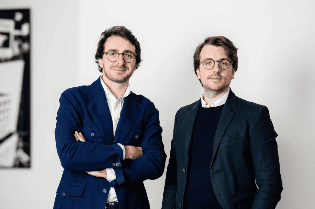 Deux hommes à lunettes posent côte à côte devant un mur blanc, l'un en veste bleu marine bras croisés, l'autre en veste sombre