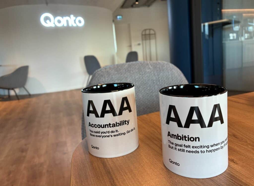 Deux mugs Qonto estampillés AAA, l’un Accountability, l’autre Ambition, sur une table en bois devant le logo lumineux