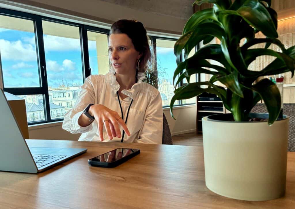 Sophie Cornay, assise à un bureau près d’une plante, gesticule en parlant, un ordinateur et un téléphone devant elle